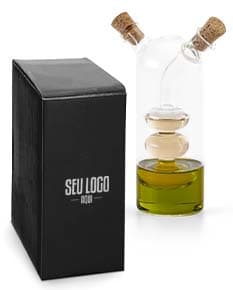 Galheteiro de vidro com três recipientes, um para azeite, outro para vinagre e um terceiro para molho de pimenta. Cada recipiente possui uma tampa de metal e um bico dosador. O galheteiro está sobre uma mesa de madeira escura.