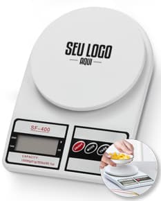 Mulher sorrindo e trabalhando em sua cozinha, com uma mesa de café da manhã com comida, xícara de café, pratos e utensílios. Ela está segurando uma balança digital para cozinha.