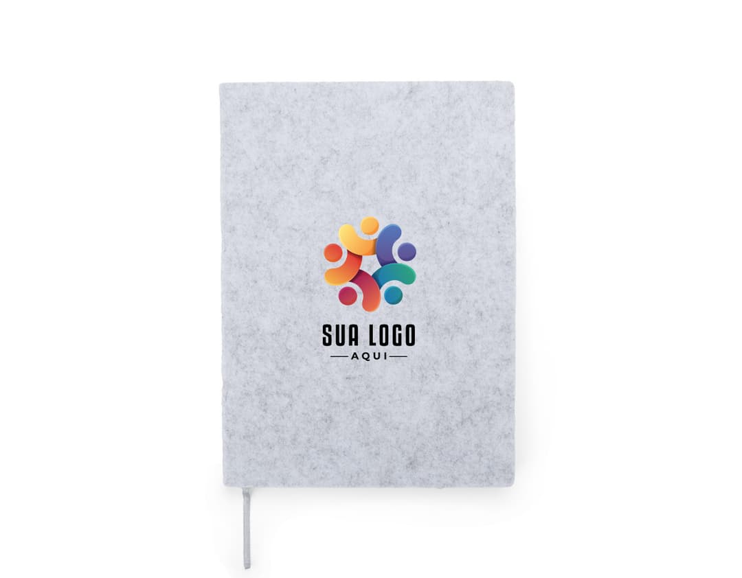 caderno A5 de feltro preto, sem pauta, aberto em uma página em branco. Ao lado, um notebook e um envelope preto. No fundo, livros e objetos de escritório.